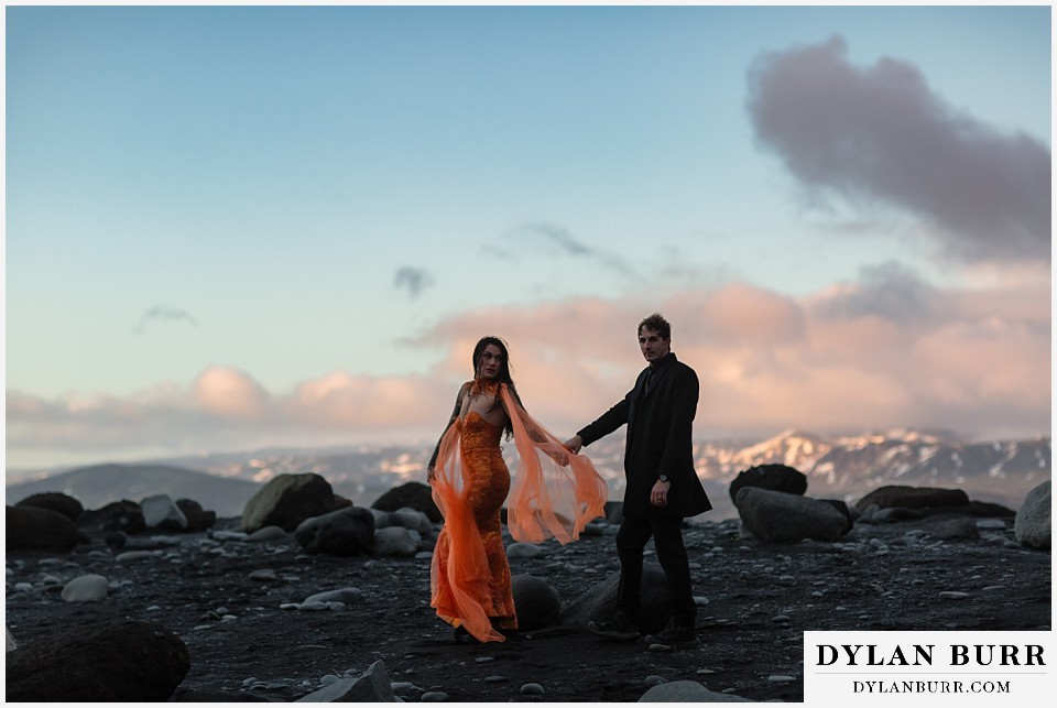 iceland elopement at reynisfjara beach orange dress walking together with mountains in the distance
