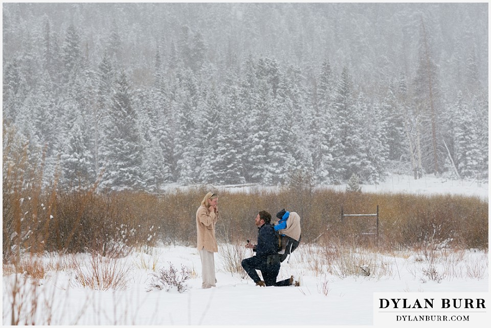 man proposes to his girlfriend with their son in a backpack behind him in the snow in Rocky Mountain National Park